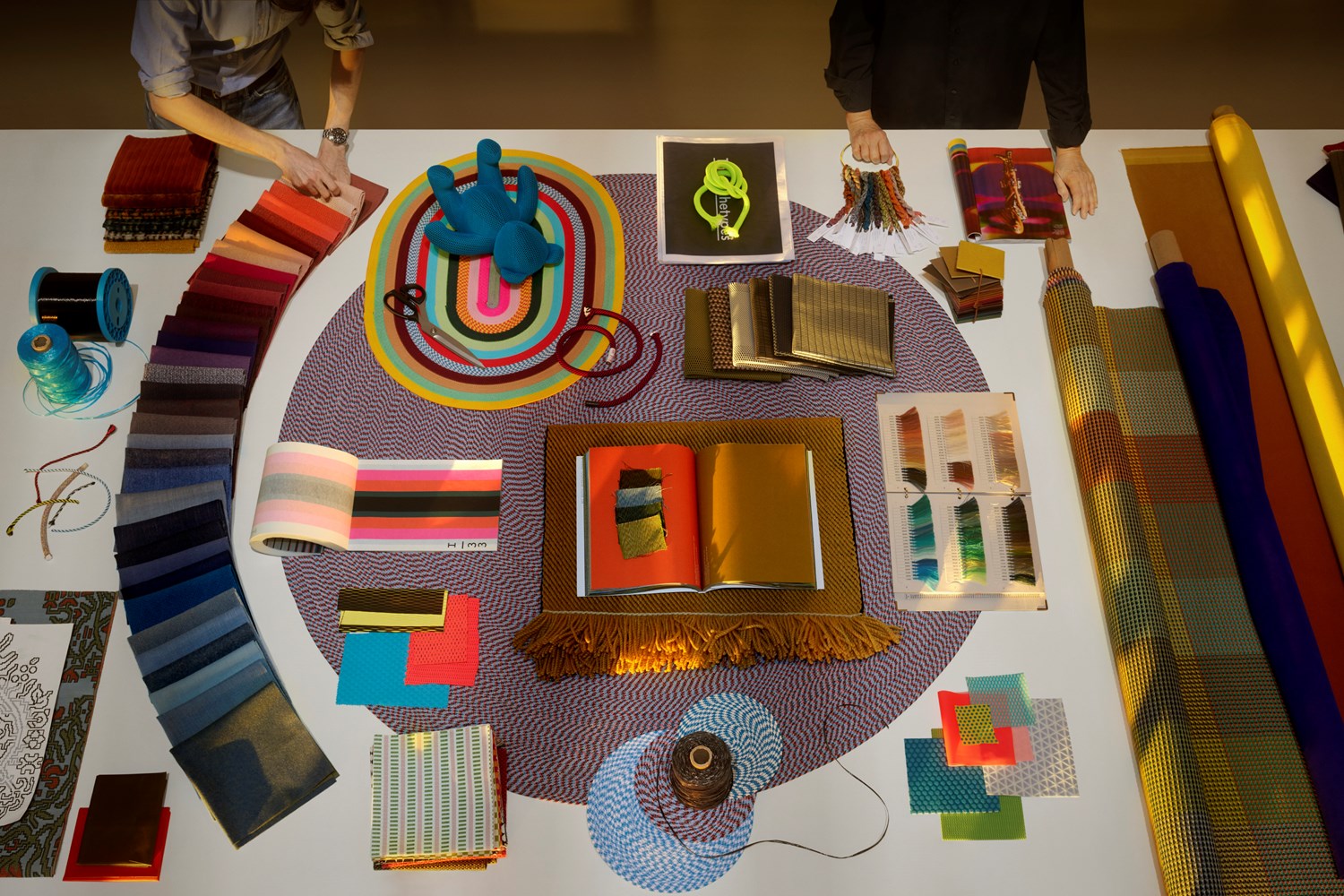 Overhead view of a design studio table with scattered textile samples and creative tools, centered around a colorful arrangement. Overhead view of a design studio table with scattered textile samples and creative tools, centered around a colorful arrangement.