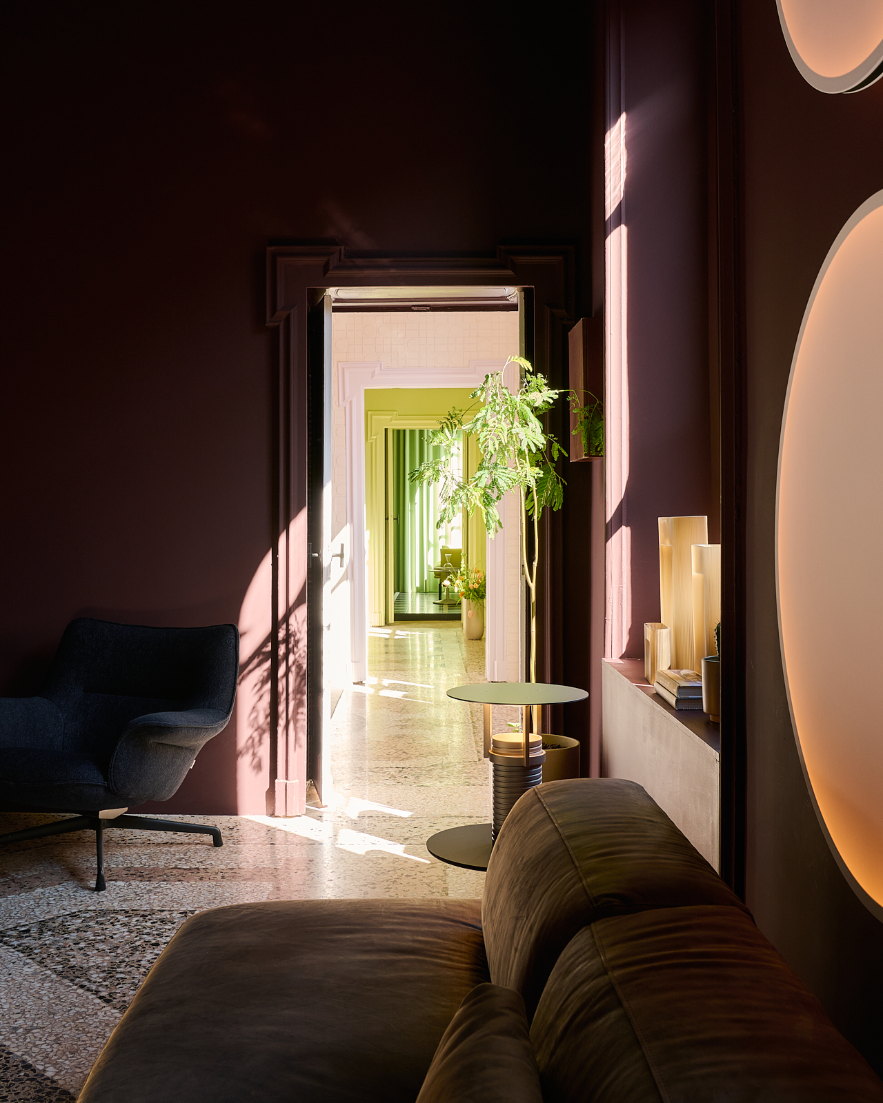 Image of Muuto's Milan Apartment. A dark dusty rose room styled with a green plant, a lounge chair, a table lamp and a brown soft sofa.