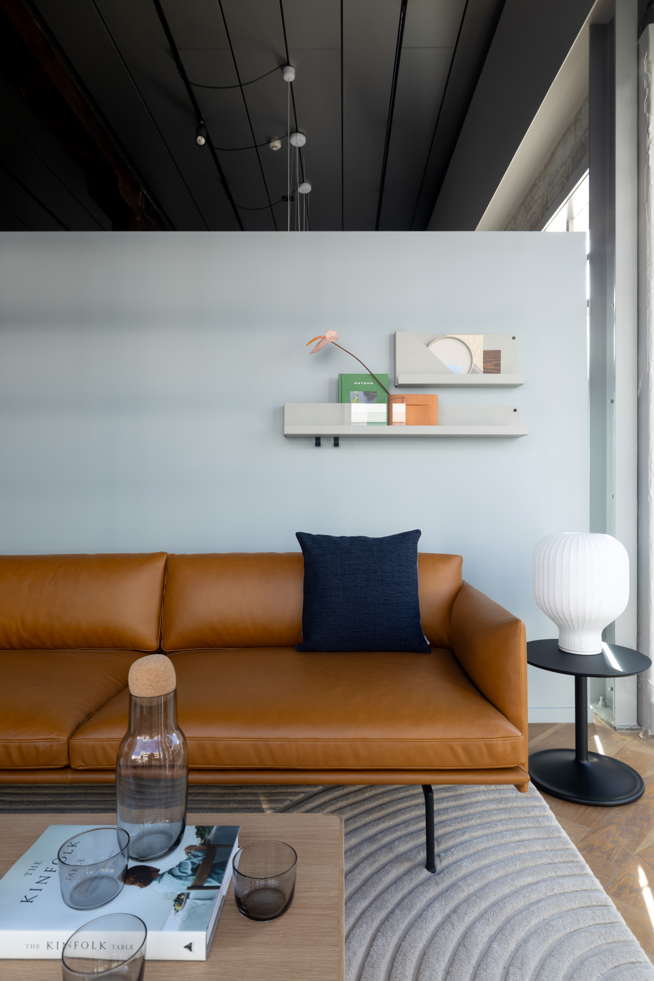 A cognac-colored leather sofa sits in a room with a wooden coffee table. A white lamp rests on a small black side table next to the sofa.