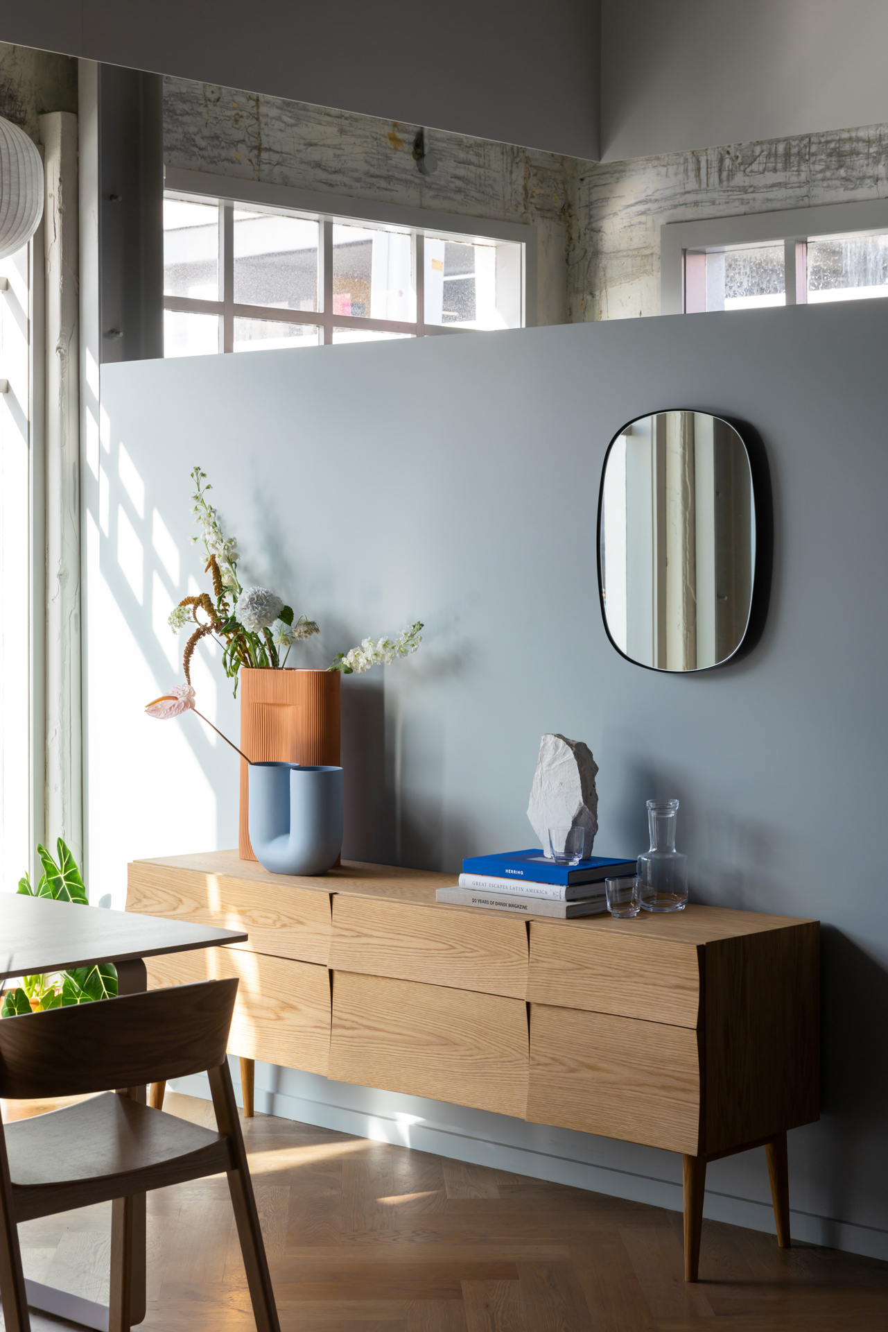 A wooden sideboard styled with sculptural vases filled with flowers, with an oval mirror hanging above the setting.