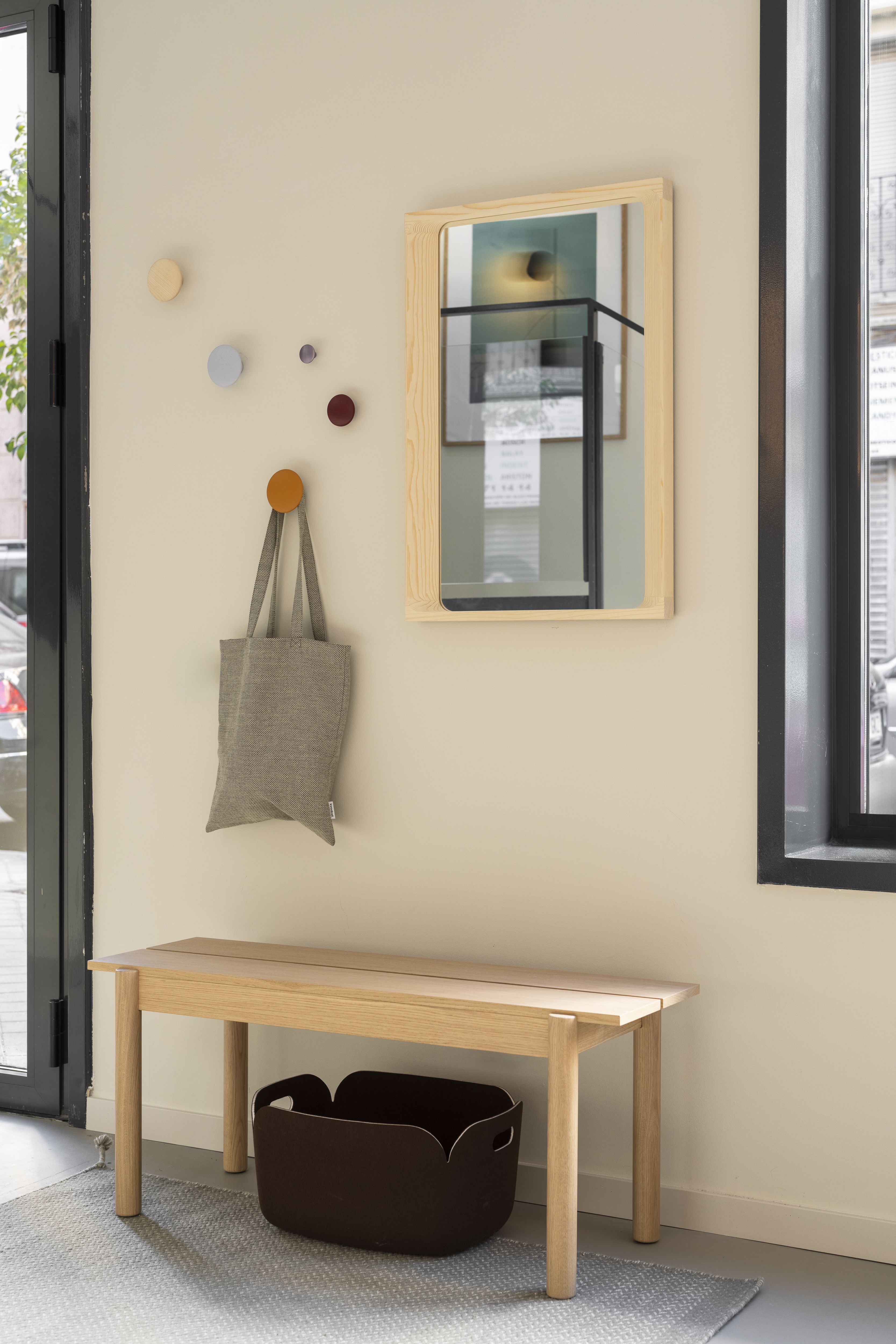 Entryway scene from the Muuto showroom in Madrid featuring the Linear Wood Bench in oak, styled with arced mirror, colorful Dots hooks  and a dark brown restore basket for a functional and welcoming setting.